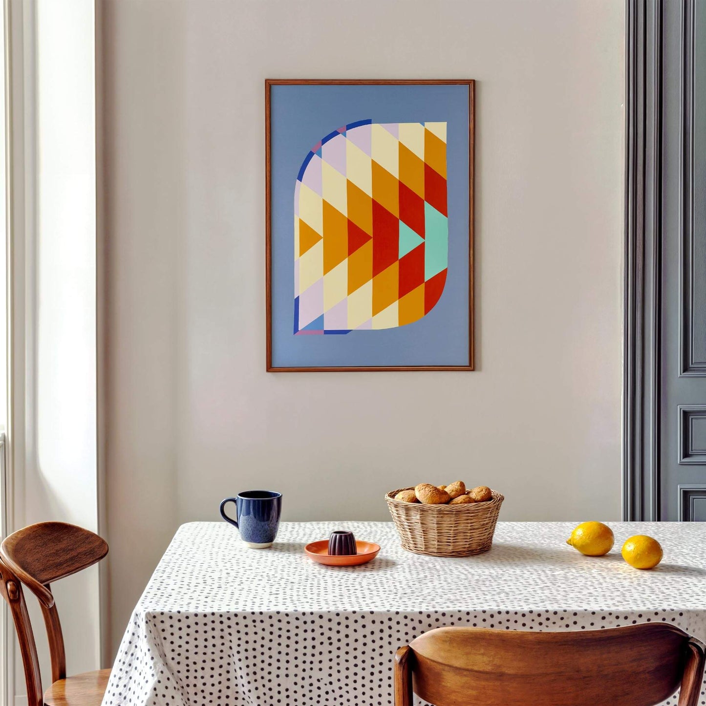 Abstract coloured print hanging above a minimal table and chairs with a cup, lemons, small manual juicer, and potatoes on top.