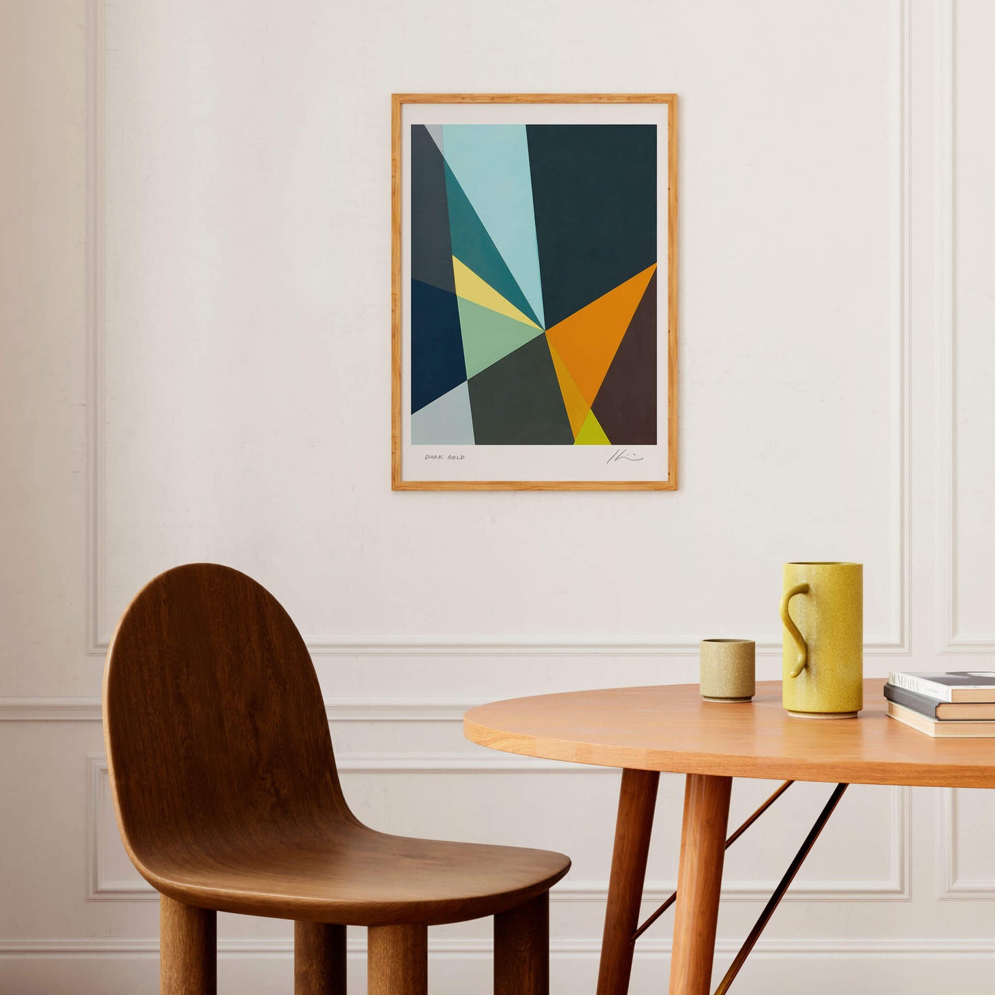 Abstract modern art print displayed above a wooden table and chair. There's a jug, a cup and some books on the table.