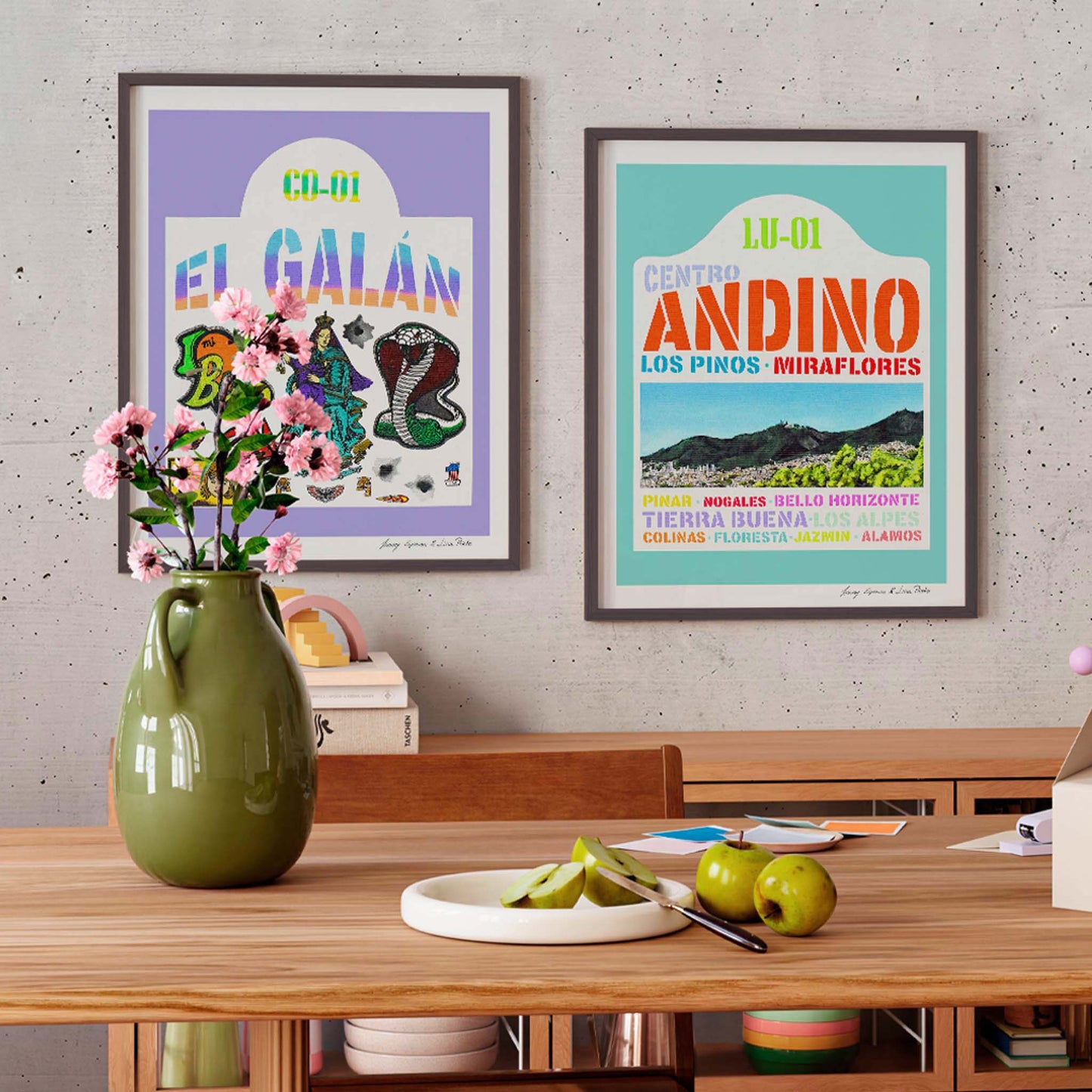 Two colourful art prints on a wall above a wooden table with a vase and apples.