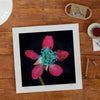 Unframed wall art print of a red flower on a wooden tabletop.