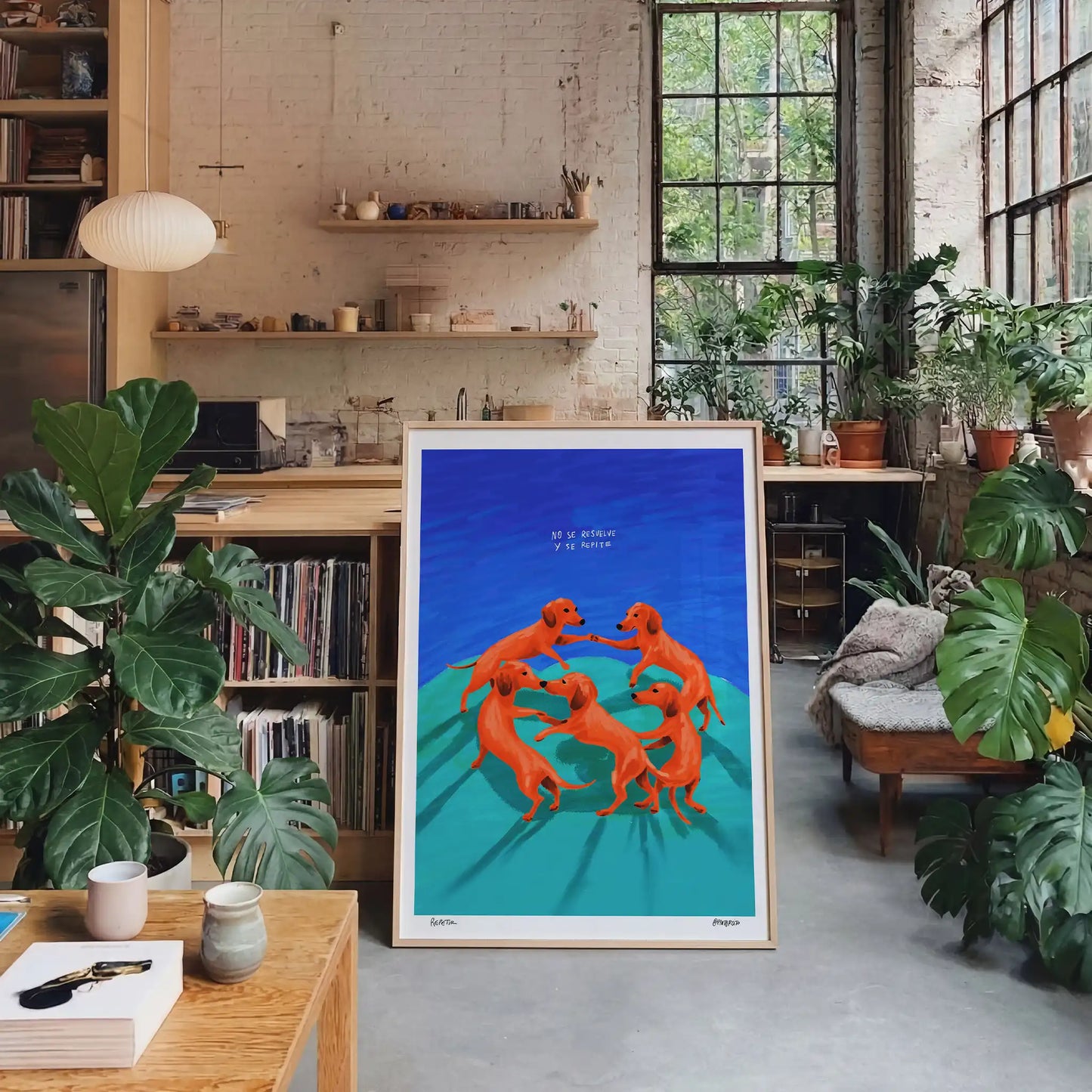 A framed art print featuring five dogs dancing in a circle on a blue and teal background, displayed in a contemporary loft filled with plants.