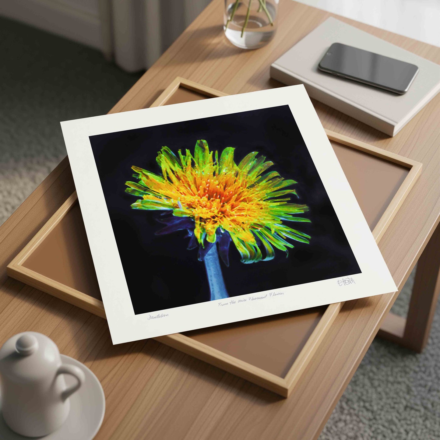Framed painting of a dandelion flower on a wooden table with a cup and phone in the background.