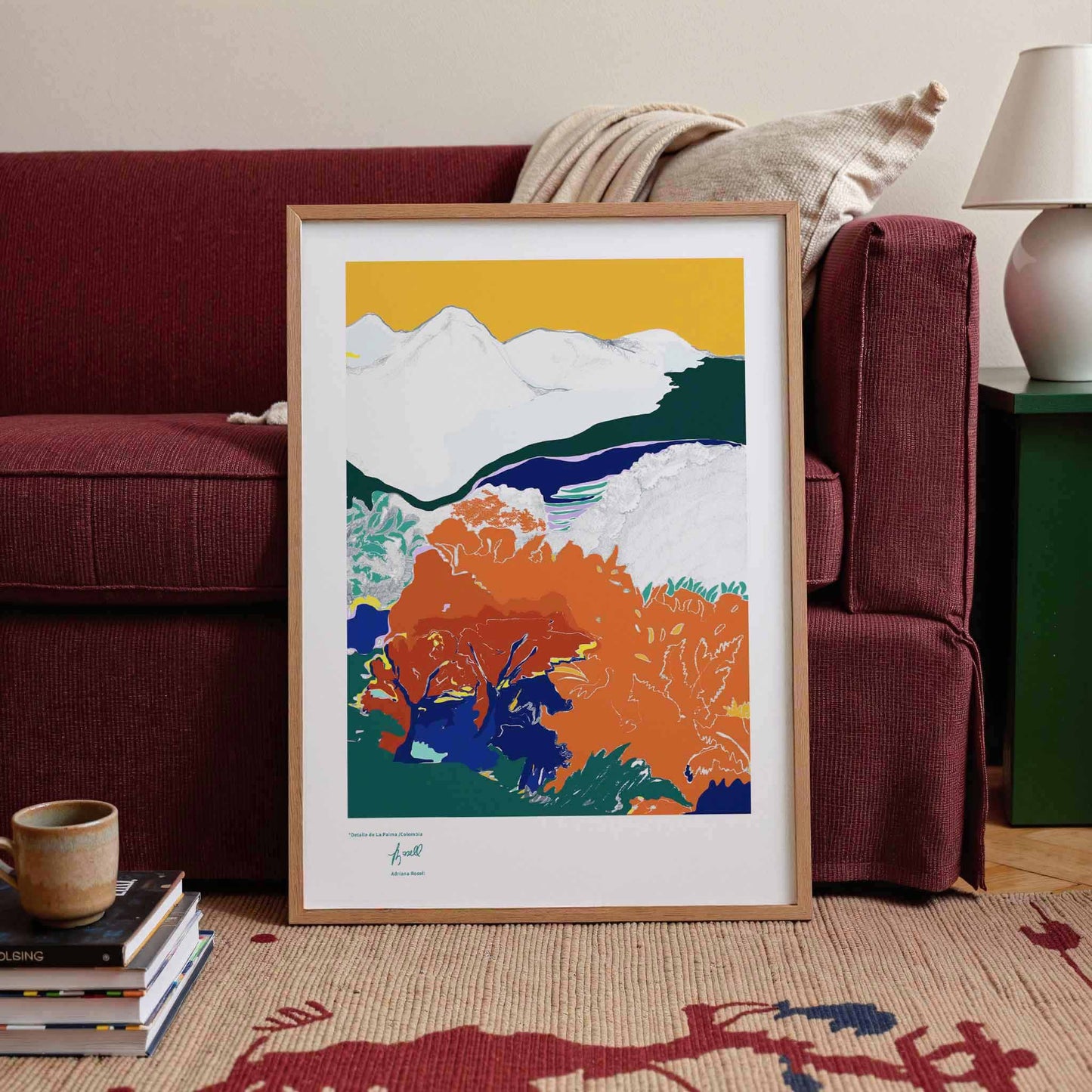 Framed landscape art print in a living room setting with a red sofa and lamp.