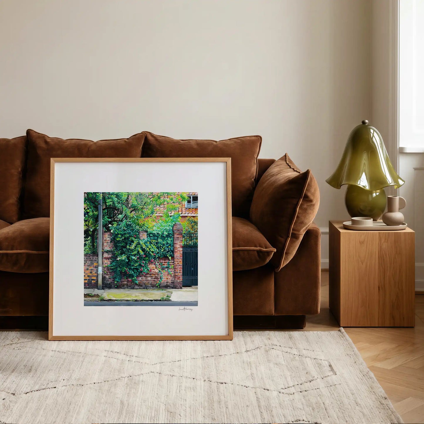 A framed art print of a brick wall with green foliage against a brown velvet sofa in a modern living room setting.