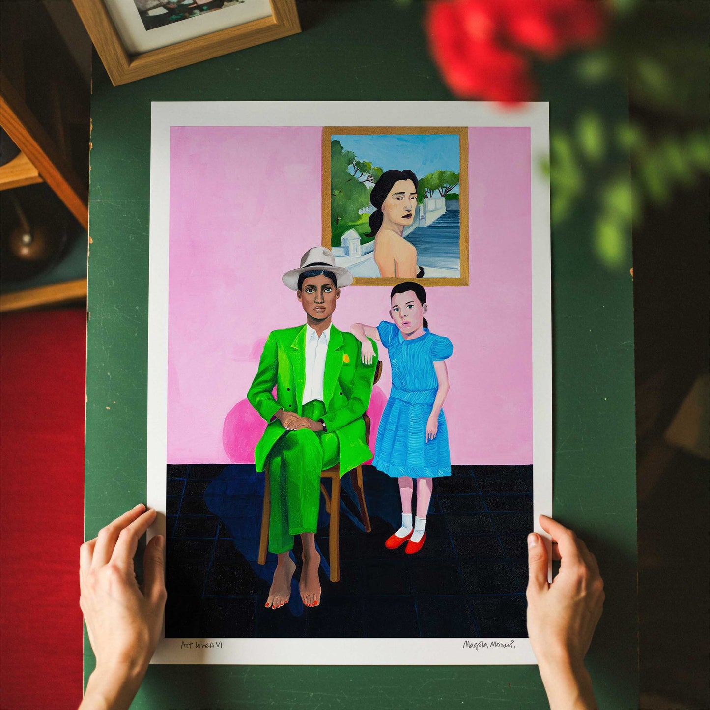 Art print of a woman in a green suit and a girl in a blue dress against a pink background, unframed and held by hands on a dark tabletop.