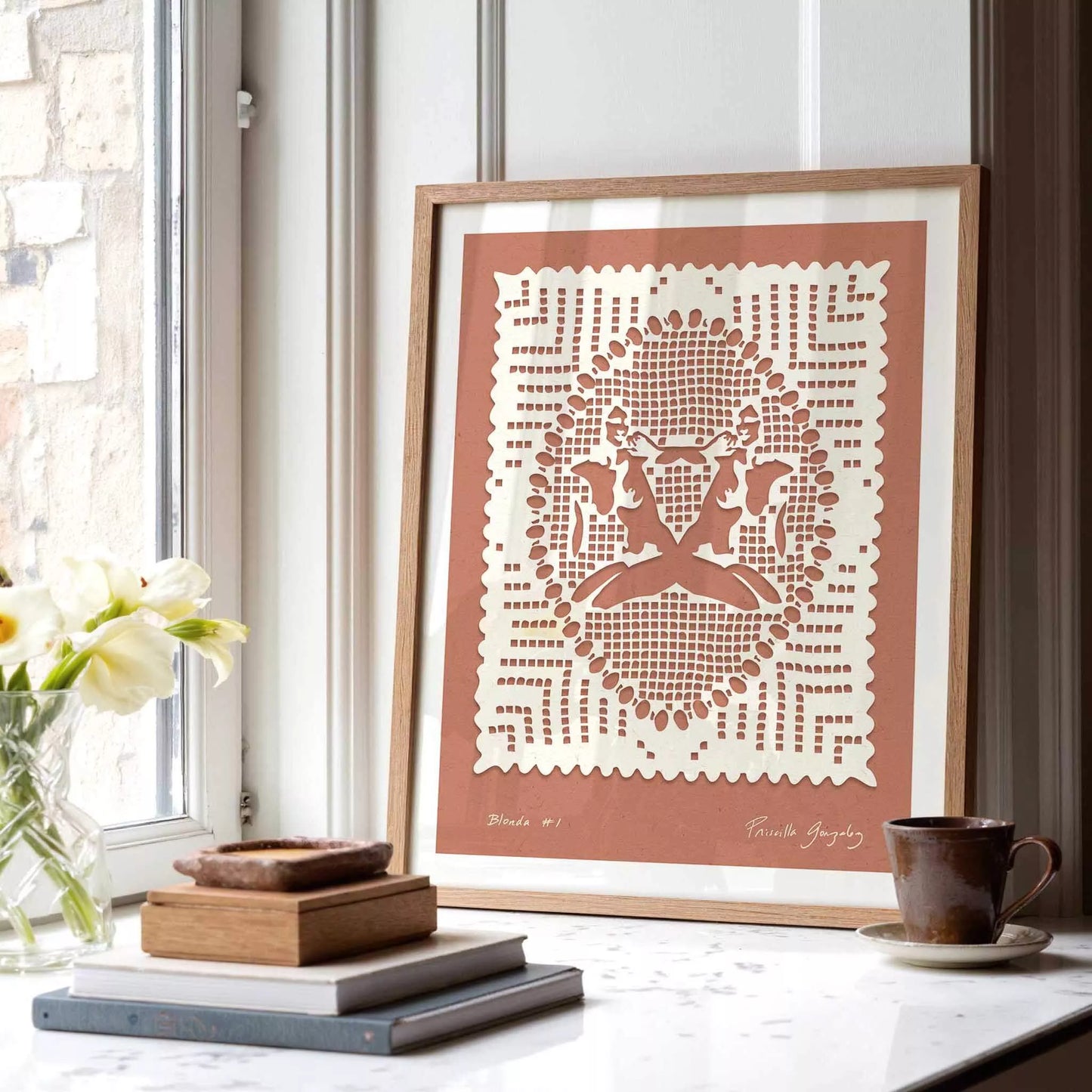 Framed papercut art of feminine figures on a windowsill with books and a cup.